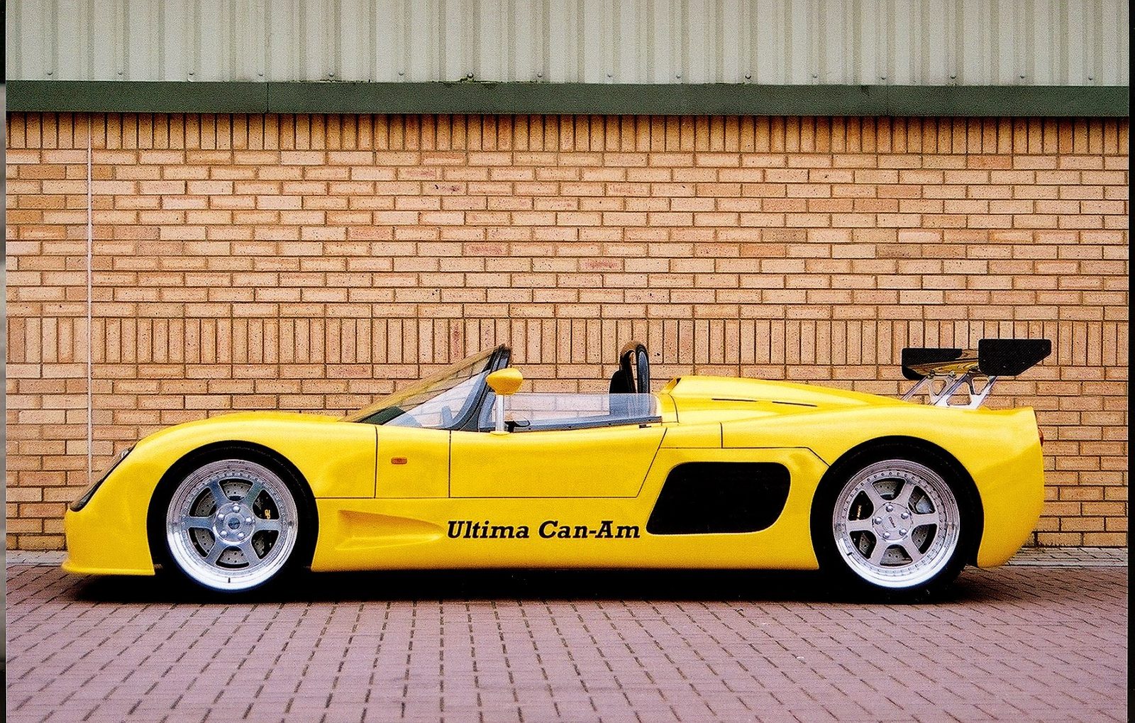 Yellow Ultima Can-Am in side profile beside a brick wall