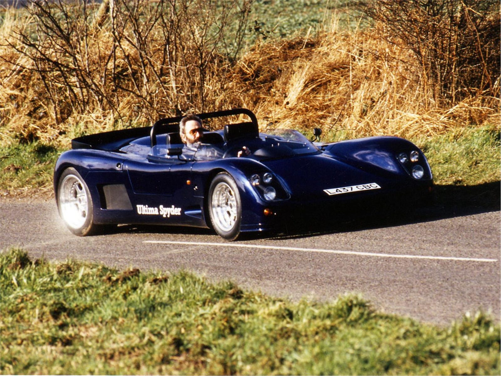 Blue Ultima Spyder driving along a road